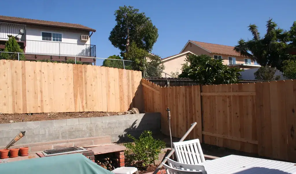 A new cedar board privacy fence around a suburban backyard, with neighboring houses and a patio table in view
